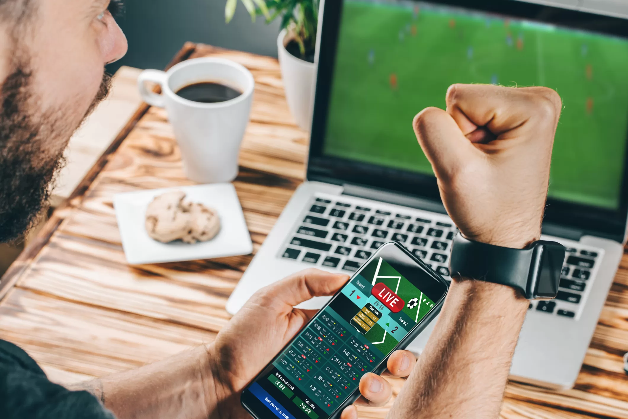 Lucky man celebrating victory after making bets using gambling mobile application on his phone. Football match online broadcast on laptop screen on the background.